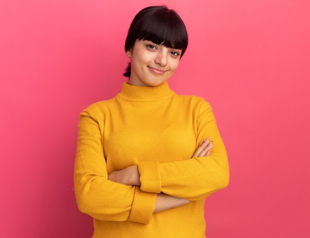 Pleased young brunette caucasian girl stands with crossed arms isolated on pink wall with copy space