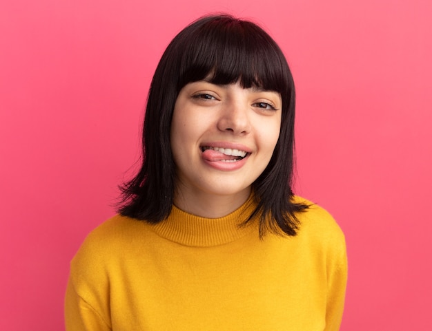 Pleased young brunette caucasian girl stucks out tongue on pink