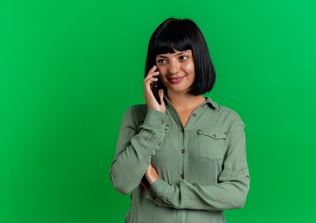 Довольная молодая брюнетка кавказская девушка разговаривает по телефону, глядя в сторону, изолированную на зеленом фоне с копией пространства
