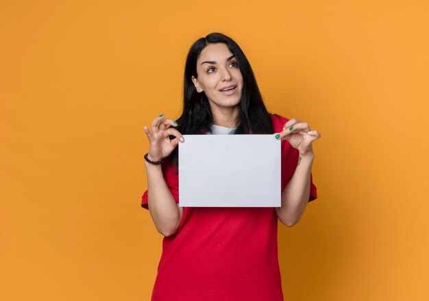 Pleased young brunette caucasian girl wearing red shirt holds blank paper sheet looking at side isolated on orange wall