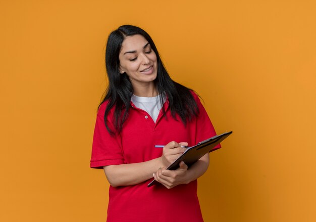 Pleased young brunette caucasian girl wearing red shirt writes with pen on clipboard isolated on orange wall