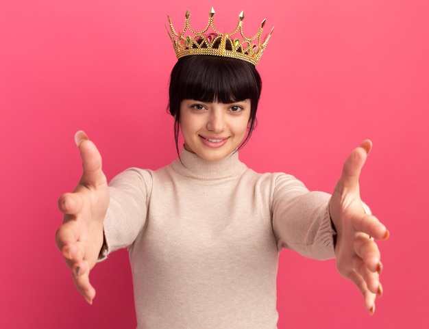 Free photo pleased young brunette caucasian girl with crown stretching out hands isolated on pink wall with copy space