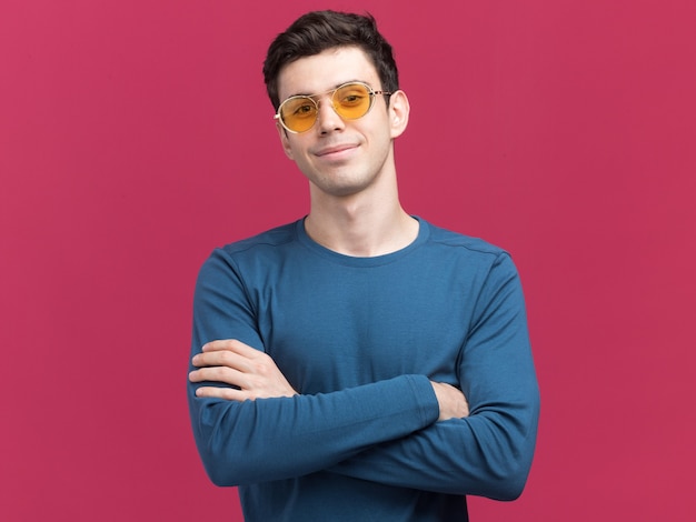 Pleased young brunette caucasian man in sun glasses standing with crossed arms isolated on pink wall with copy space