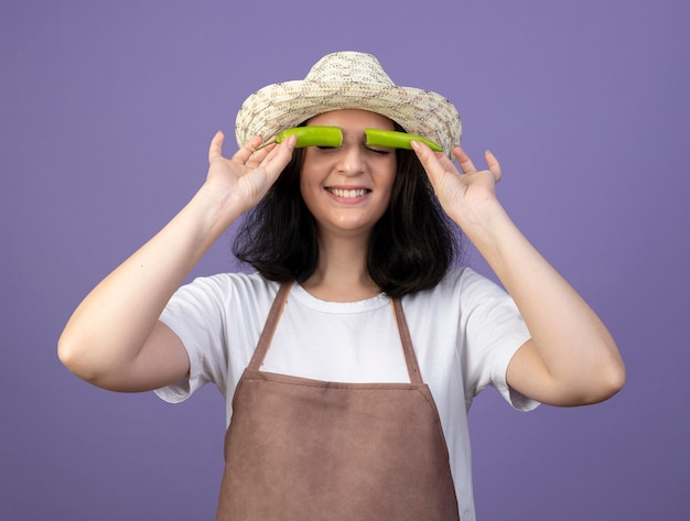 無料写真 ガーデニング帽子をかぶって制服を着た若いブルネットの女性の庭師は、紫色の壁に分離された唐辛子の半分で目を覆っています