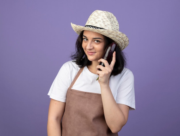 Pleased young brunette female gardener in uniform wearing gardening hat holds eggplant isolated on purple wall