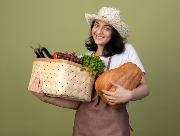 Free photo pleased young brunette female gardener in uniform wearing gardening hat holds vegetable basket and pumpkin isolated on olive green wall