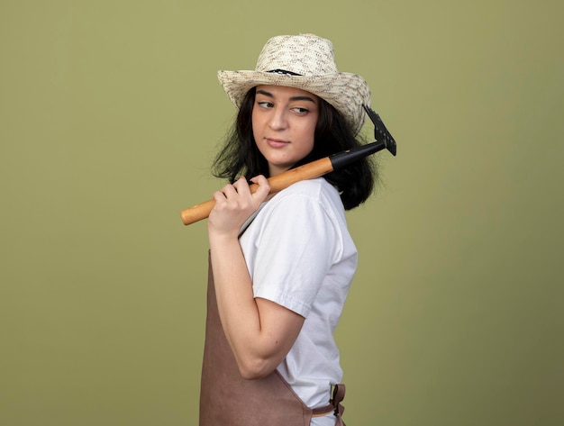 Pleased young brunette female gardener in uniform wearing gardening hat stands sideways