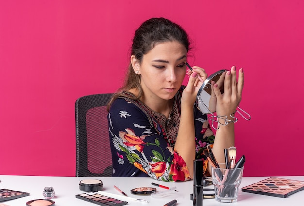 Pleased young brunette girl sitting at table with makeup tools applying eyeshadow with makeup brush holding and looking at mirror isolated on pink wall with copy space