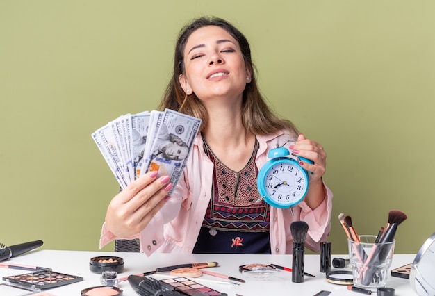 Pleased young brunette girl sitting at table with makeup tools holding money and alarm clock  isolated on olive green wall with copy space