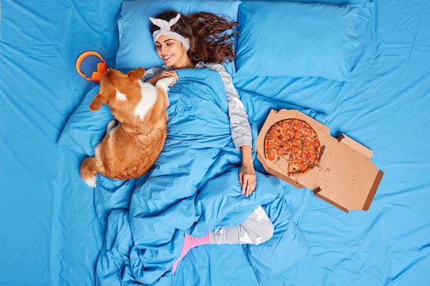 満足している若いブルネットの女性は、ベッドから出るのが面倒である快適なパジャマに身を包んだ犬と遊ぶおいしいピザを食べるすべての仕事を忘れてよく眠った後、お気に入りのペットと一緒にリラックス