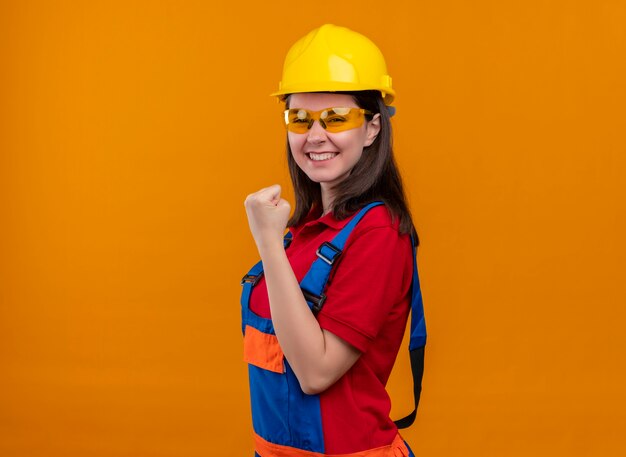 Довольная молодая девушка-строитель в защитных очках поднимает кулак и стоит боком на изолированном оранжевом фоне с копией пространства