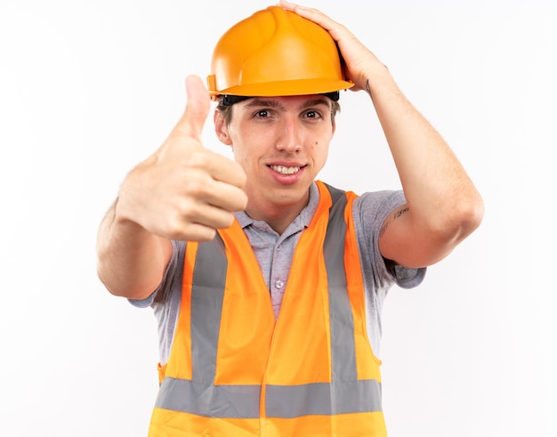 Довольный молодой строитель в форме показывает палец вверх, положив руку на голову, изолированную на белой стене