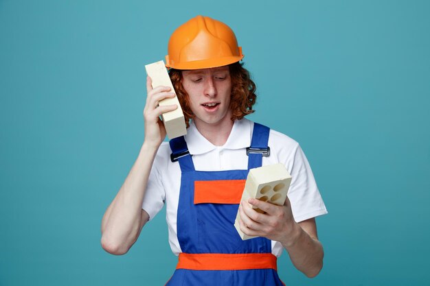 Pleased young builder man in uniform holding bricks around ear isolated on blue background