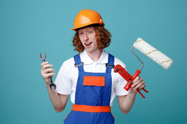 Pleased young builder man in uniform holding construction tools isolated on blue background