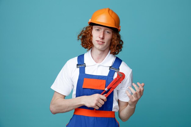 Pleased young builder man in uniform holding gas wrench isolated on blue background