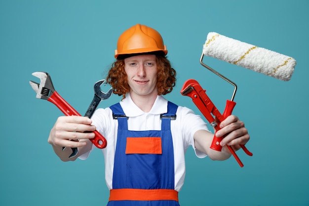 Pleased young builder man in uniform holding out construction tools at camera isolated on blue background