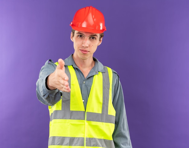 Довольный молодой строитель в униформе, протягивая руку, изолированную на синей стене