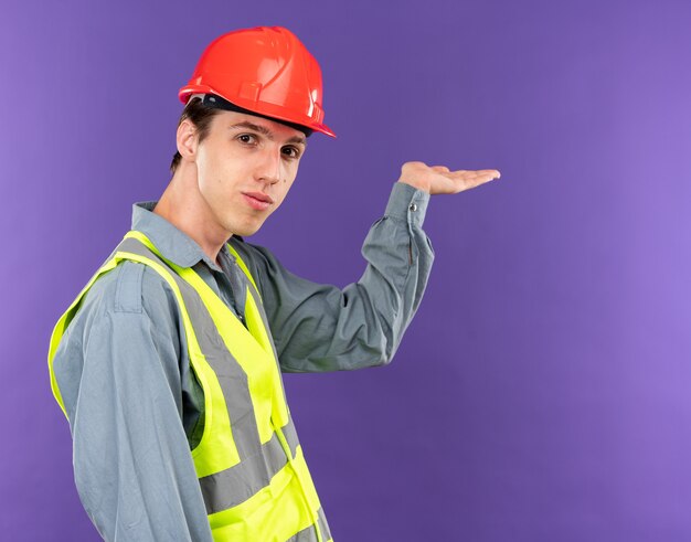 Pleased  young builder man in uniform points at behind isolated on blue wall with copy space