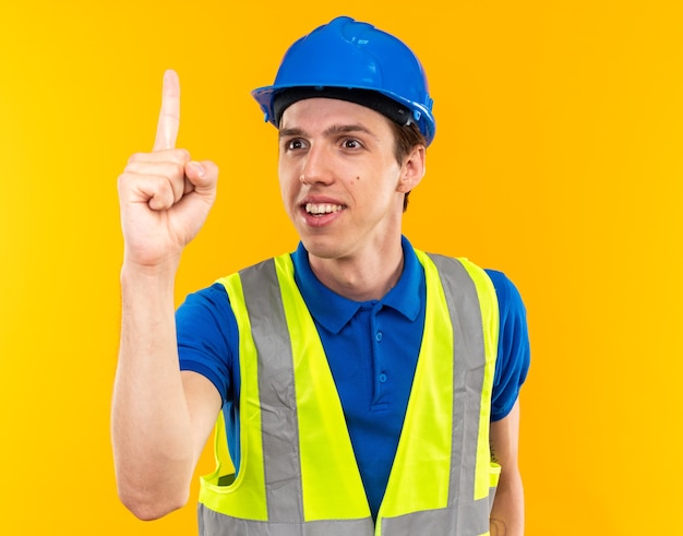 Довольный молодой человек-строитель в униформе, показывающий один на желтой стене