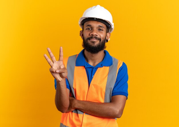 довольный молодой человек-строитель в униформе со защитным шлемом, жестикулирующий тремя пальцами, изолированными на оранжевой стене с копией пространства