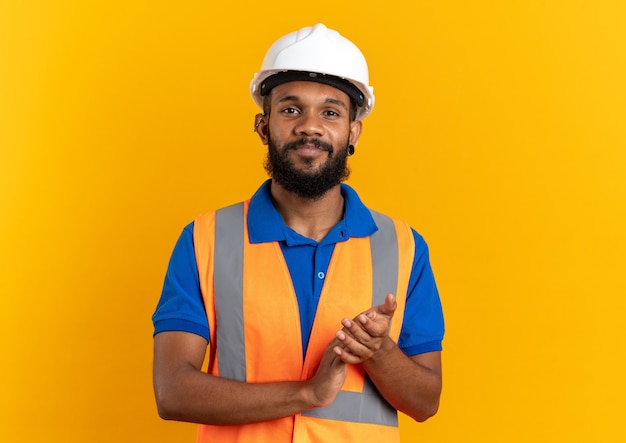 Pleased young builder man in uniform with safety helmet holding hands together isolated on orange wall with copy space