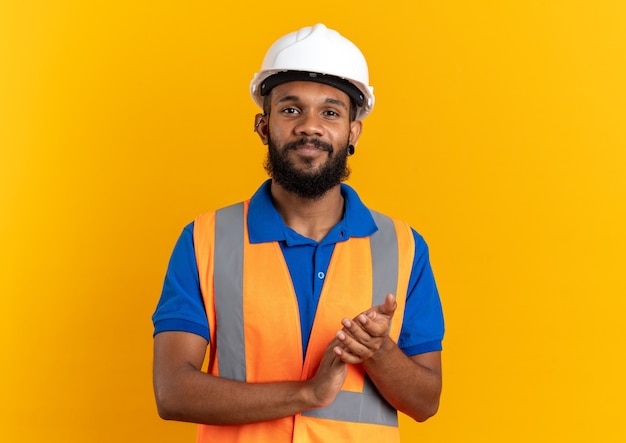 pleased young builder man in uniform with safety helmet holding hands together isolated on orange wall with copy space