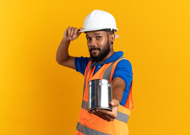 pleased young builder man in uniform with safety helmet holding oil paint isolated on orange wall with copy space