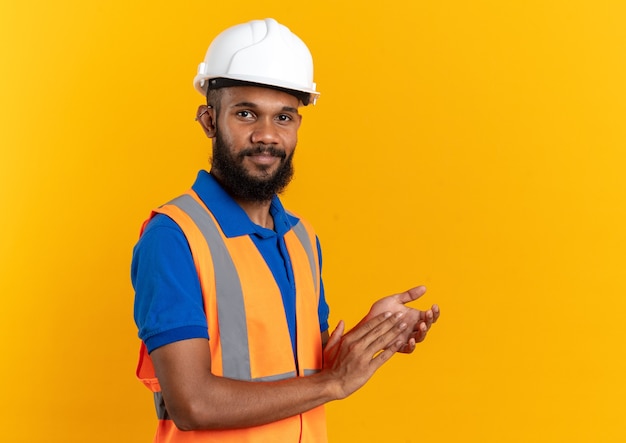 Pleased young builder man in uniform with safety helmet standing sideways holding hands together isolated on orange wall with copy space