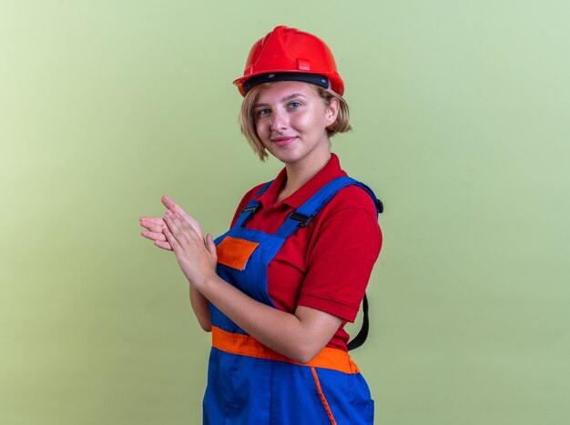 pleased young builder woman in uniform holding hands together isolated on olive green wall