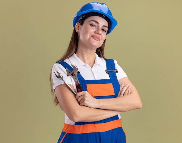 Pleased young builder woman in uniform holding open-end wrench crossing hands isolated on olive green wall