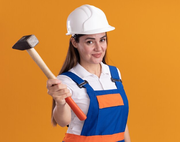 Pleased young builder woman in uniform holding out hammer at camera isolated on orange wall