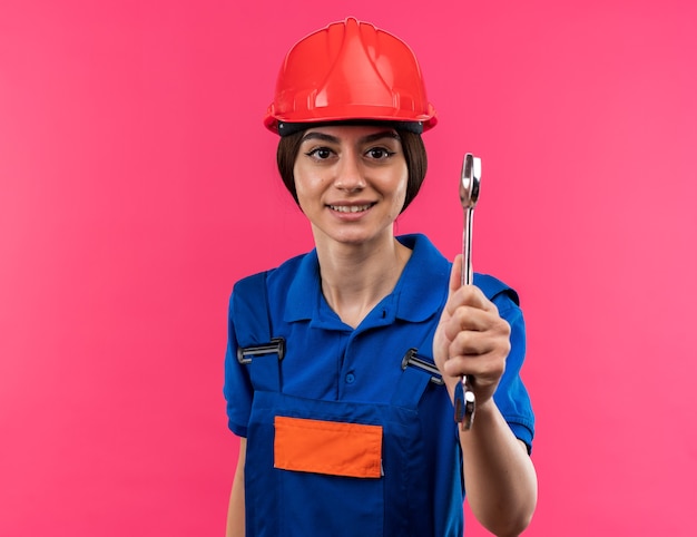 Pleased young builder woman in uniform holding out open-end wrench isolated on pink wall