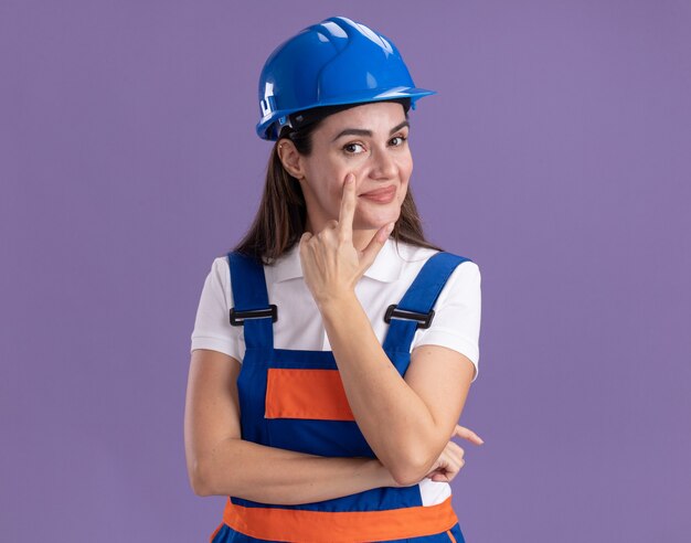 Lieta giovane donna costruttore in uniforme tirato giù la palpebra isolata sulla parete viola