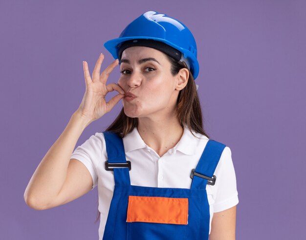 Pleased young builder woman in uniform showing delicious gesture isolated on purple wall