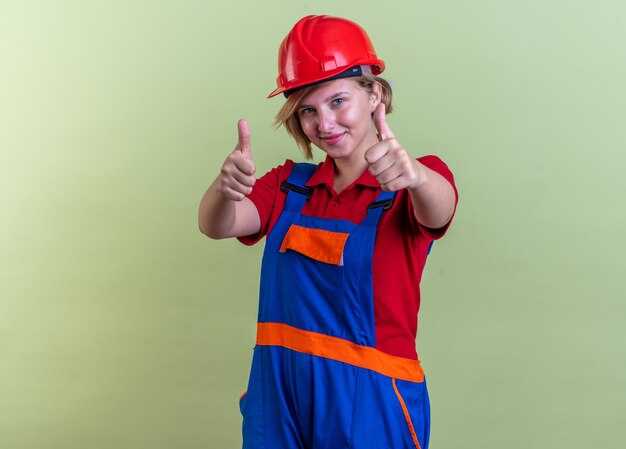 pleased young builder woman in uniform showing thumbs up isolated on olive green wall