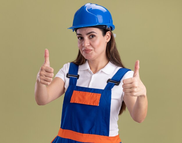 Lieta giovane donna costruttore in uniforme che mostra i pollici in su isolato sulla parete verde oliva