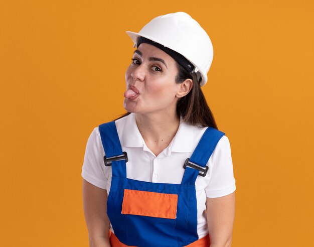 オレンジ色の壁に分離された舌を示す制服を着た若いビルダーの女性を喜ばせる