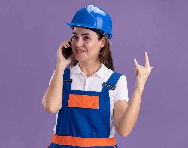 Pleased young builder woman in uniform speaks on phone and showing goat gesture isolated on purple wall