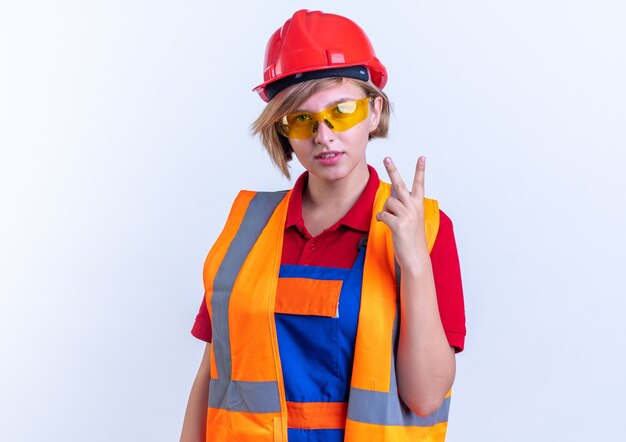 pleased young builder woman in uniform with glasses showing peace gesture isolated on white wall