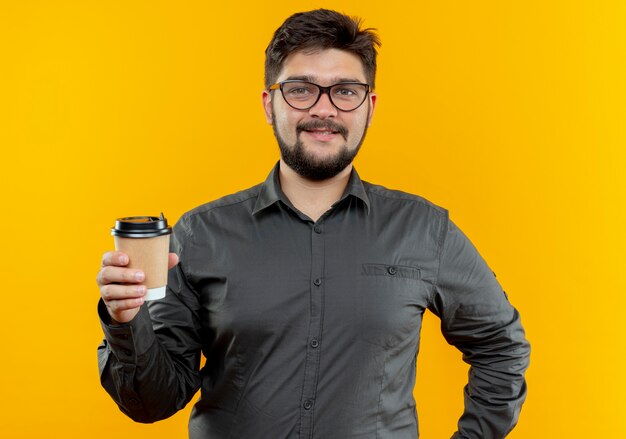 Довольный молодой бизнесмен в очках держит чашку кофе, изолированную на желтой стене