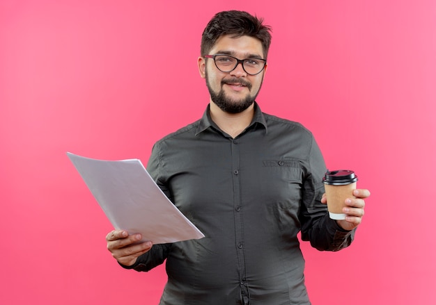 Pleased young businessman wearing glasses holding cup of coffee and  paper isolated on pink wall