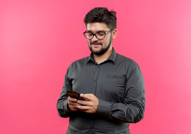 Pleased young businessman wearing glasses holding and looking at phone isolated on pink wall