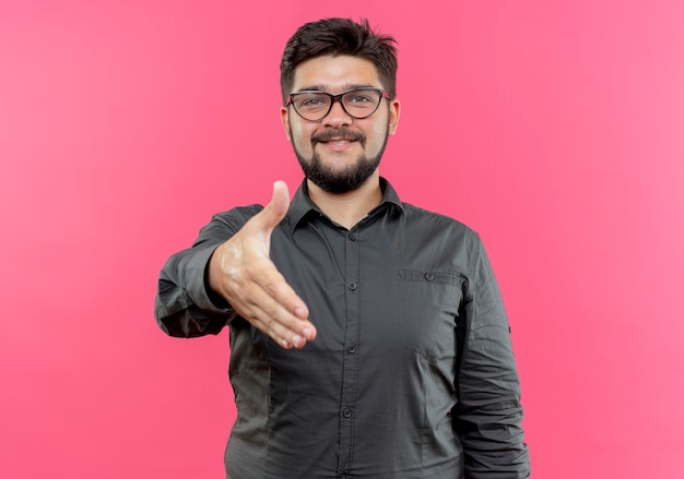 Pleased young businessman wearing glasses holding out hand isolated on pink wall