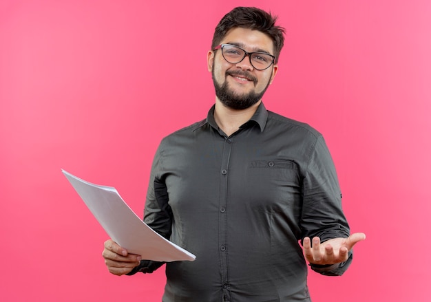 Pleased young businessman wearing glasses holding paper and holding out hand  isolated on pink wall