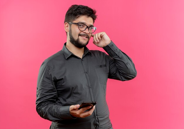 Pleased young businessman wearing glasses holding phone putting finger on ear isolated on pink wall