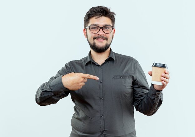 Доволен молодой бизнесмен в очках Холдинг и указывает на чашку кофе, изолированные на белом фоне