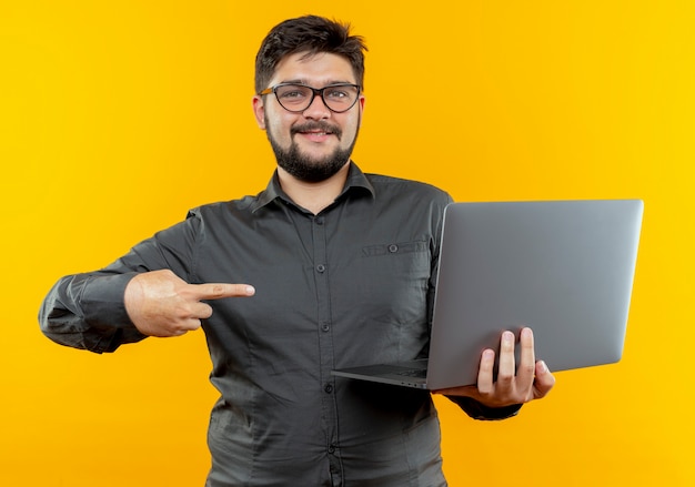 Доволен молодой бизнесмен в очках держит и указывает на ноутбук, изолированные на желтом фоне