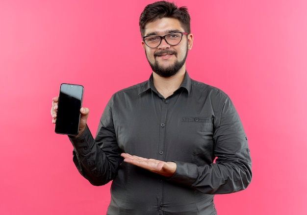 pleased young businessman wearing glasses holding and points with hand at phone
