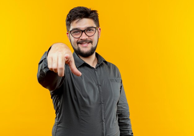Pleased young businessman wearing glasses points at camera isolated on yellow background