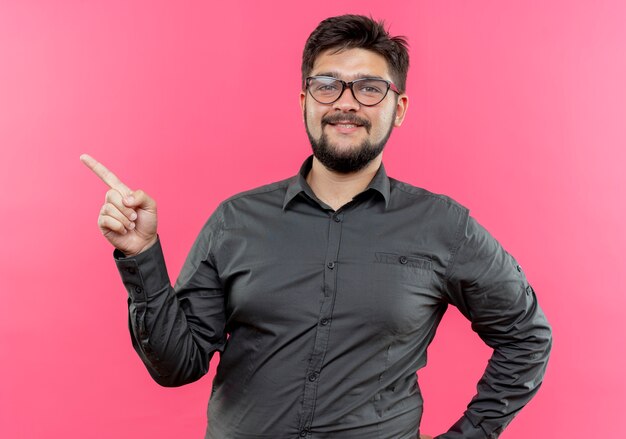 pleased young businessman wearing glasses points at side isolated on pink with copy space